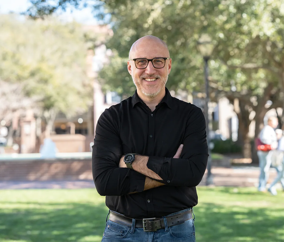 A smiling man with glasses stands outdoors on a college campus with his arms crossed, wearing a black button-down shirt and jeans, with trees, a fountain, and blurred people in the background.