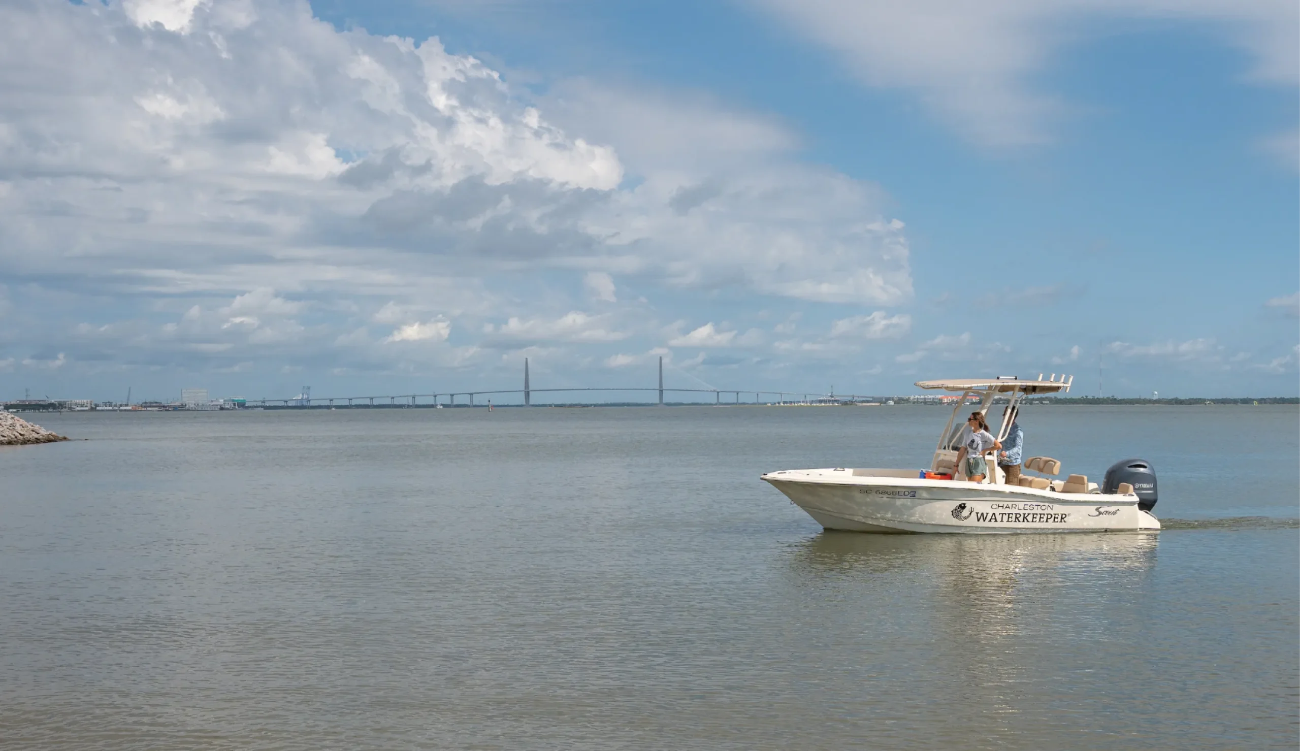 A Charleston Waterkeeper research boat floats on a wide, calm body of water with a long bridge visible in the distance beneath a partly cloudy sky.