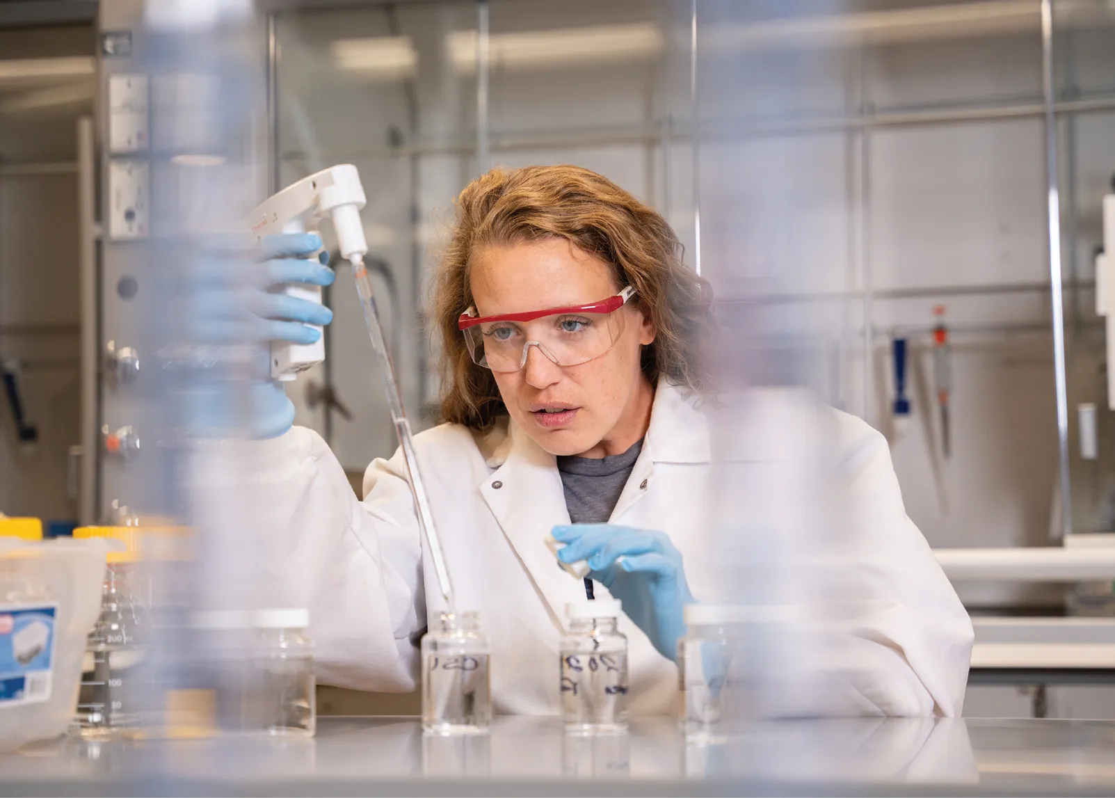 A researcher wearing safety goggles and gloves uses a pipette in a laboratory, carefully transferring liquid into small labeled glass vials on a lab bench.