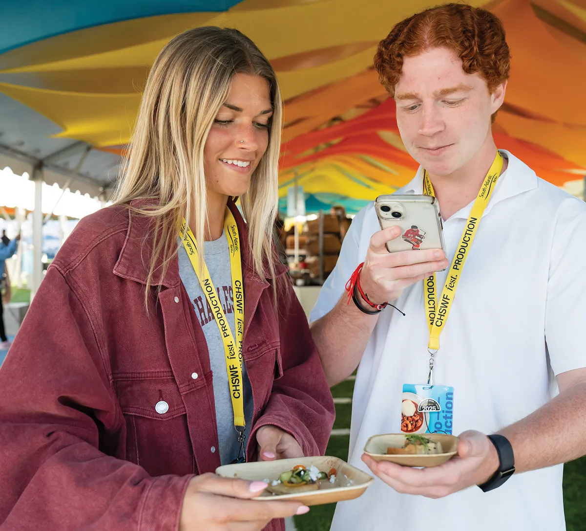 Two Charleston Wine & Food festival production members wearing yellow lanyards and holding small plates of gourmet appetizers.