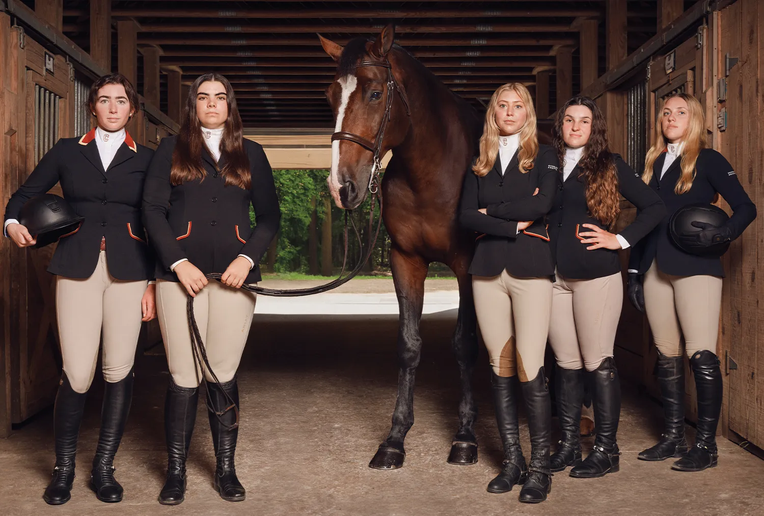 Five female equestrian team members in formal riding attire posing with a brown horse inside a wooden stable at the College of Charleston.