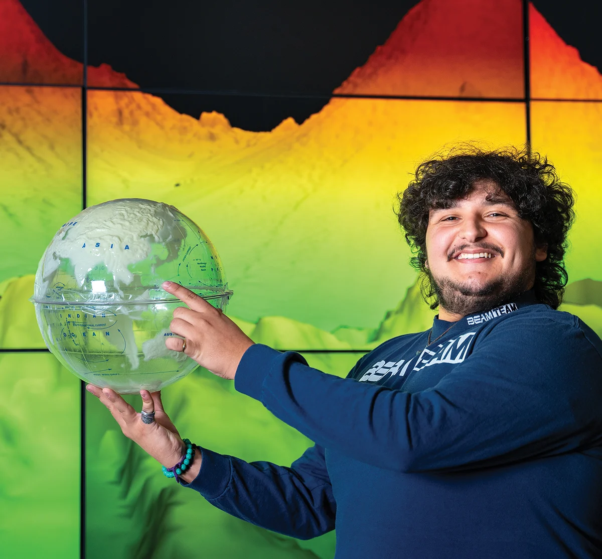 A smiling student holding a transparent 3D globe model in front of a vibrant green and orange topographic map display.