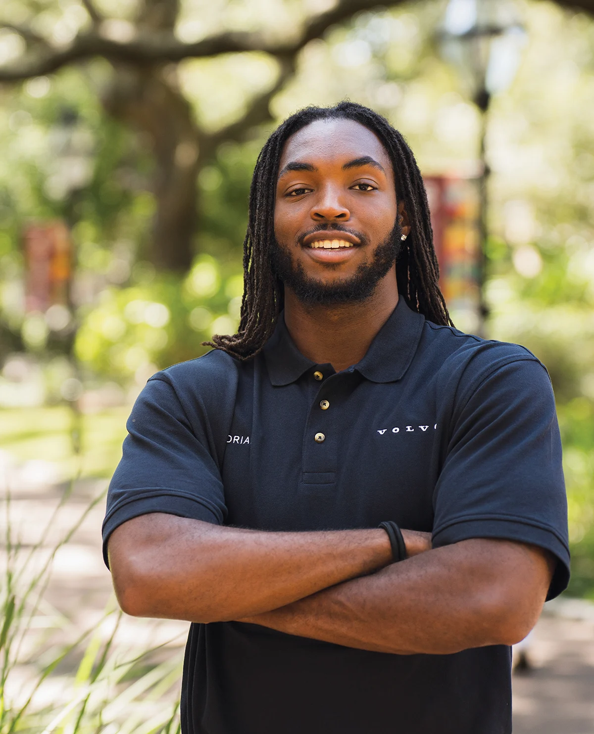 Portrait of a smiling young man with long dreadlocks wearing a black Volvo branded polo shirt in a lush, outdoor park setting.