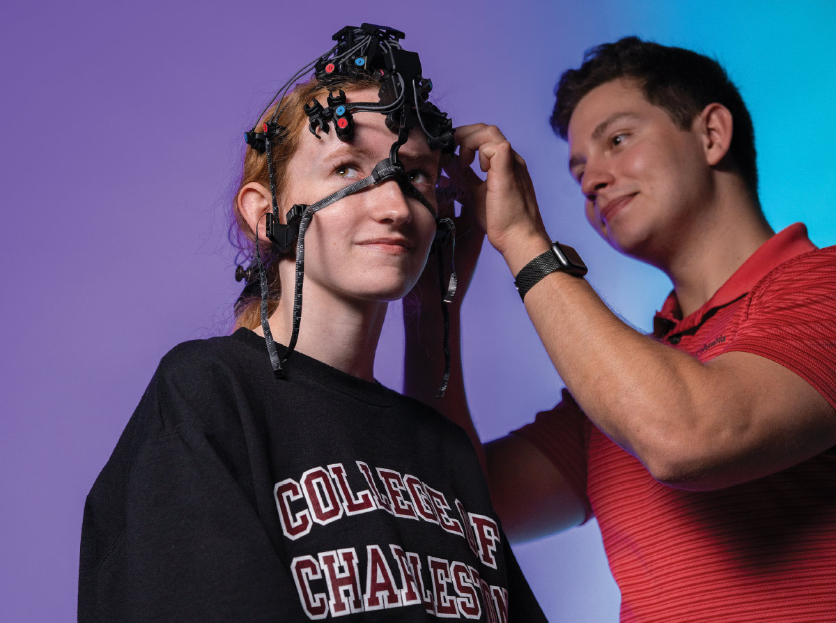 A man in a red shirt adjusts fNIRS on a woman wearing a College of Charleston sweatshirt.