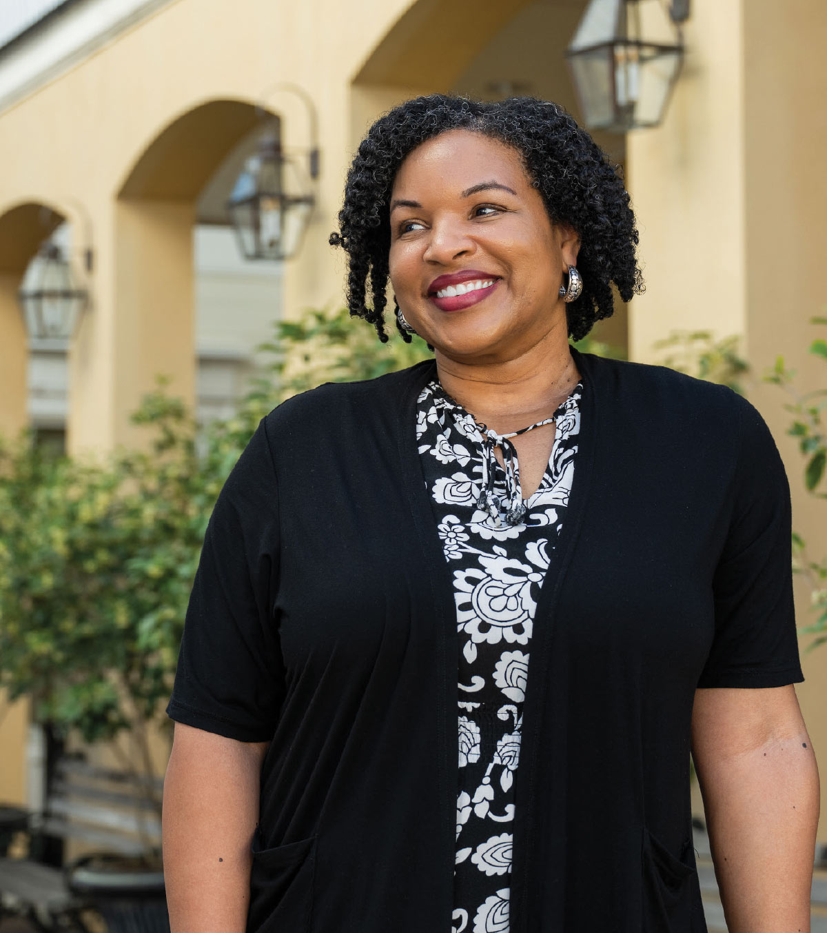 Portrait of a smiling woman with dark curly hair wearing a black cardigan and floral shirt.