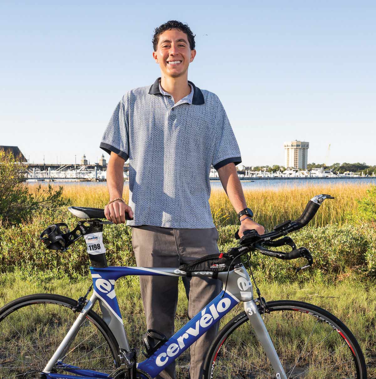A smiling man in a polo shirt stands with a silver and blue triathlon bike near a coastal marsh.