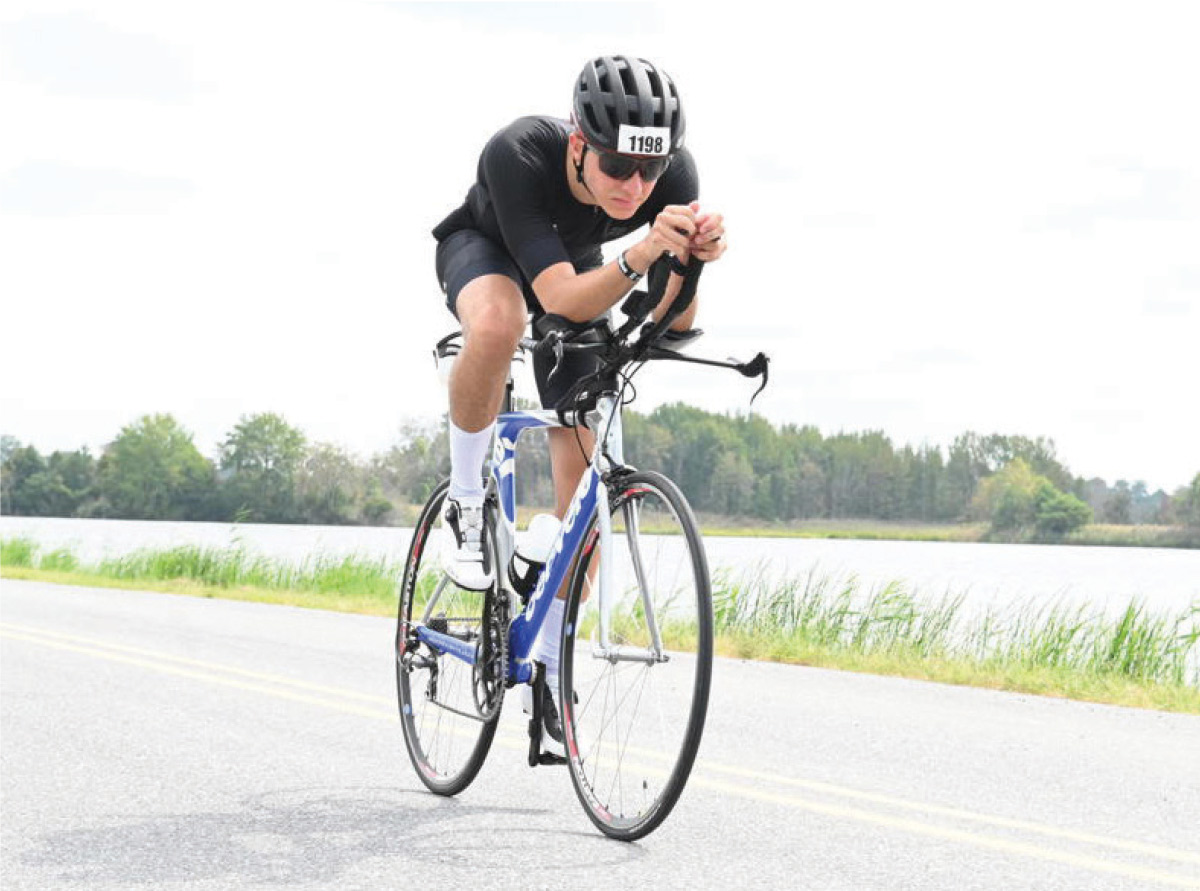 A cyclist in a black kit and helmet rides a triathlon bike on a road alongside a body of water.