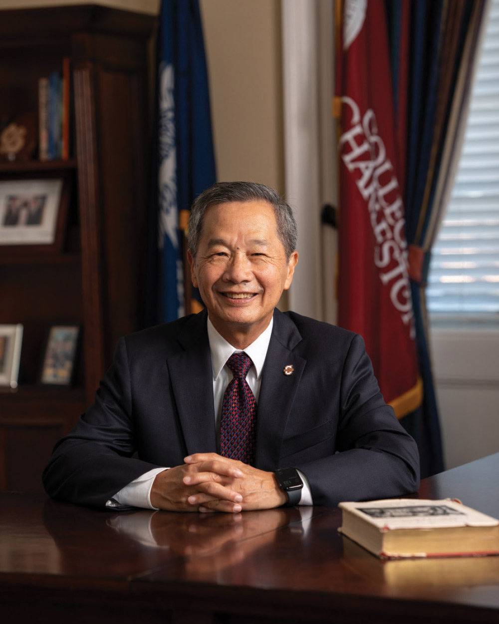 Portrait of a smiling man in a suit and tie sitting at a desk with his hands clasped, with a College of Charleston flag in the background.