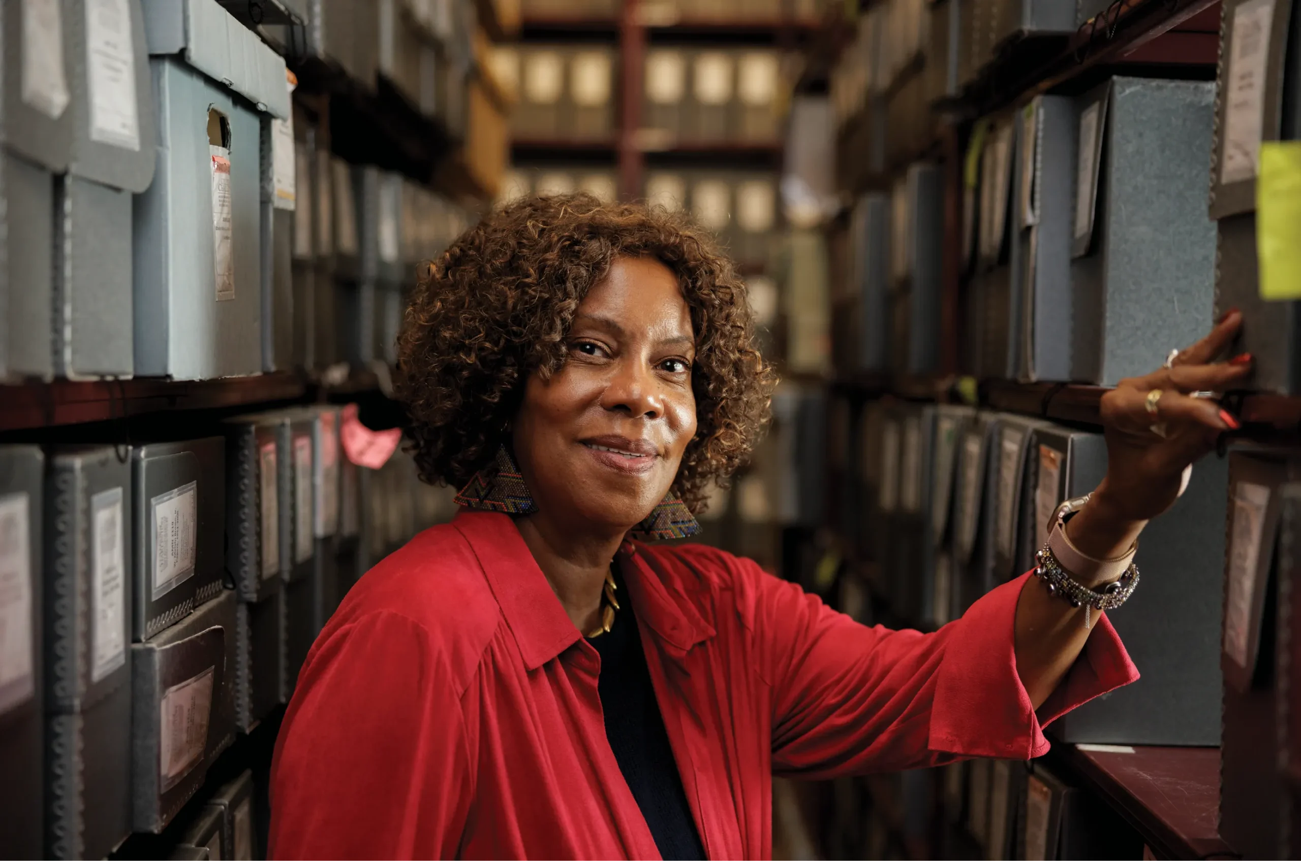 A woman stands in a climate-controlled archive aisle, smiling while resting one hand on a shelf of labeled archival boxes surrounding her on both sides.