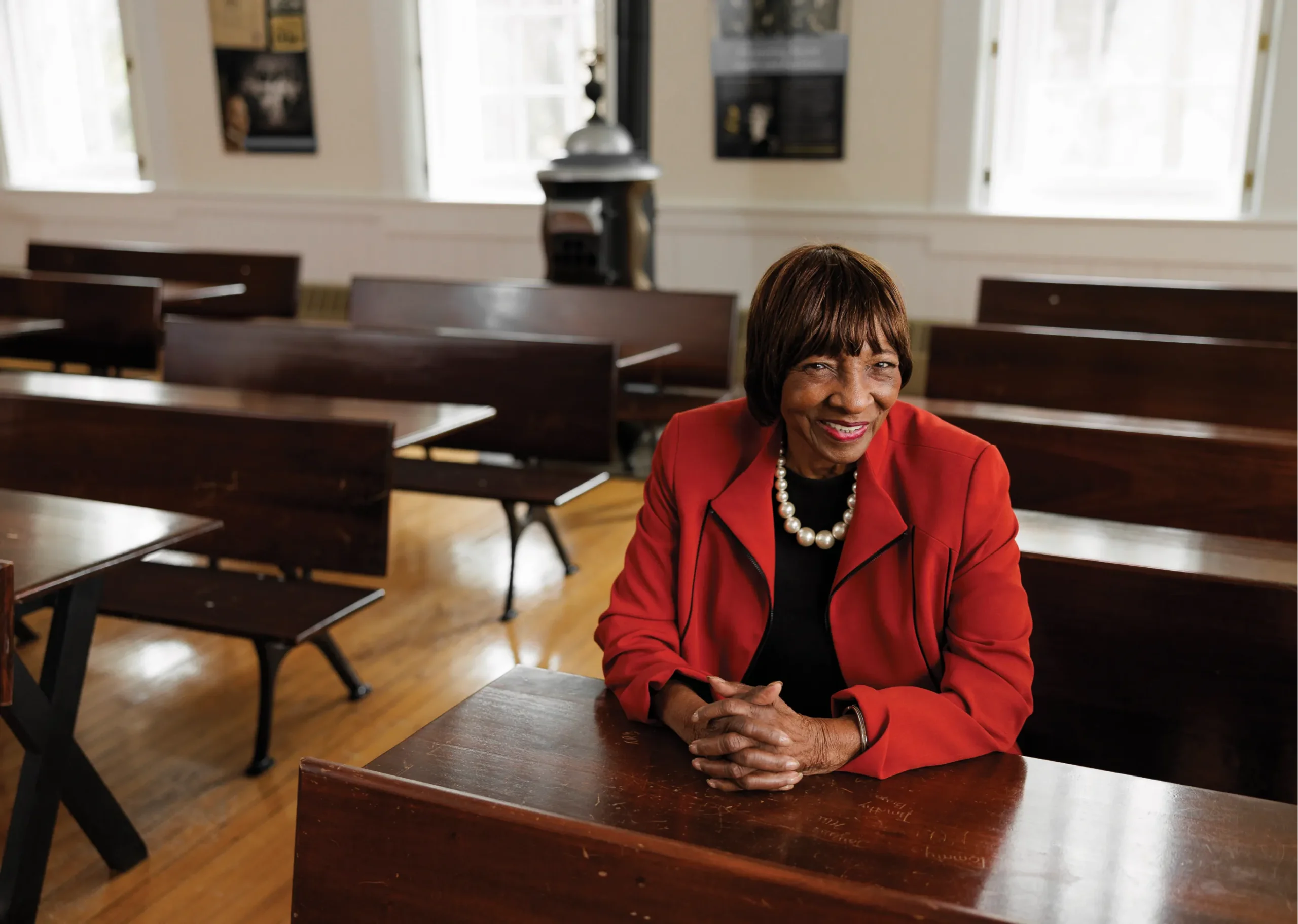 An elderly woman wearing a red jacket and pearl necklace sits at a wooden desk inside a historic classroom with rows of wooden benches, smiling warmly toward the camera.