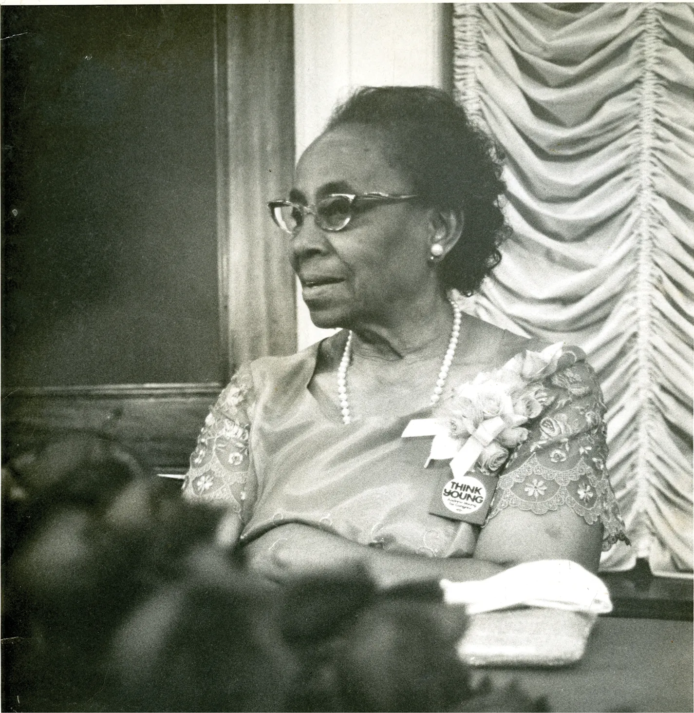 Black-and-white portrait of an older African American woman wearing glasses, a corsage, and a pearl necklace, seated at a table during a formal event.