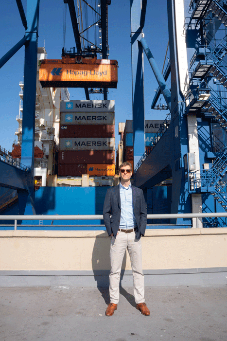 Man in sunglasses and blazer standing at a shipyard as a crane lifts an orange Hapag-Lloyd container above Maersk stacks.