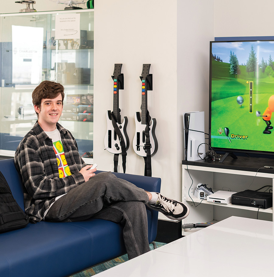 David Koch on a blue couch holding a controller, surrounded by video game consoles and guitar peripherals.