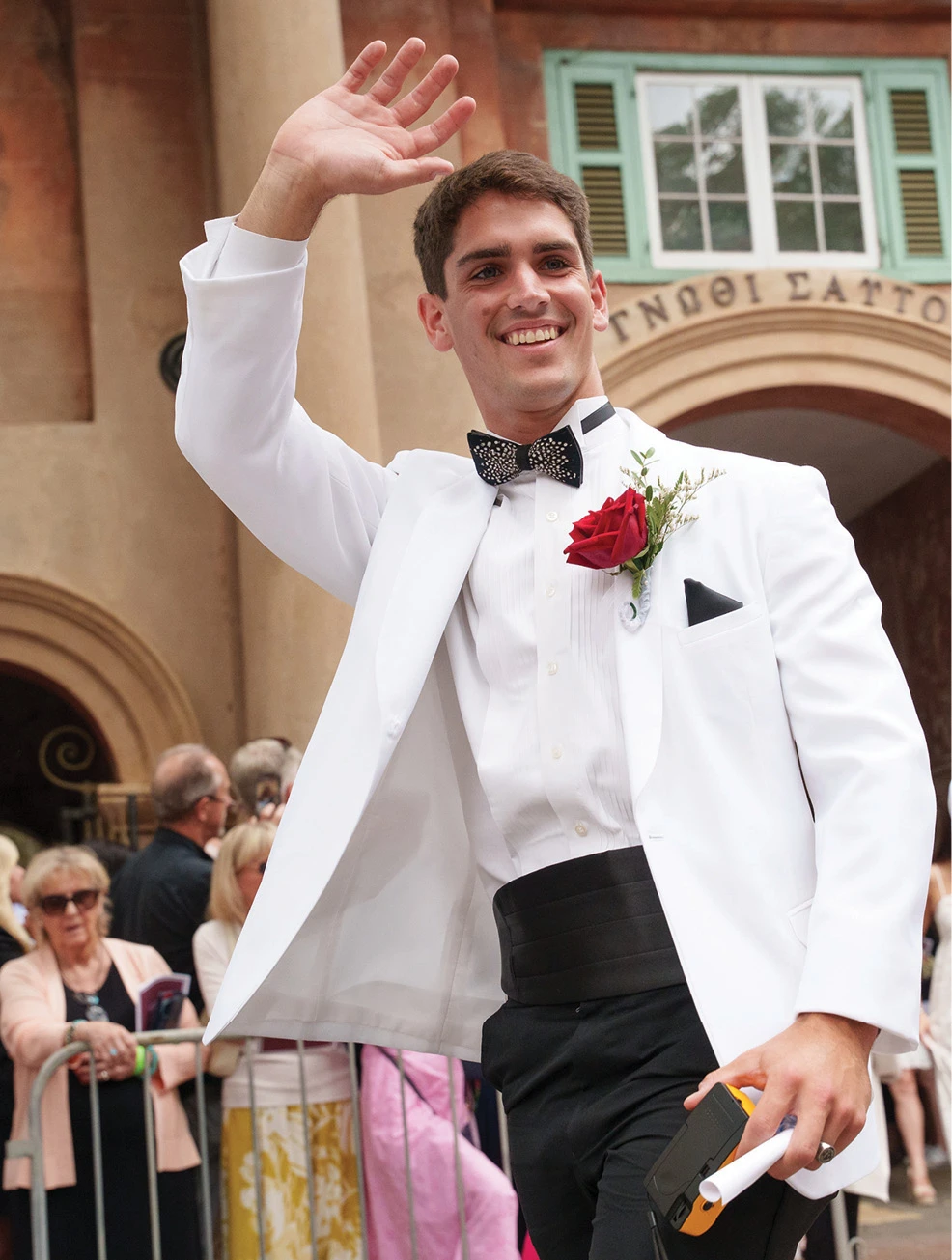 Student in a white dinner jacket waving during a campus commencement ceremony.