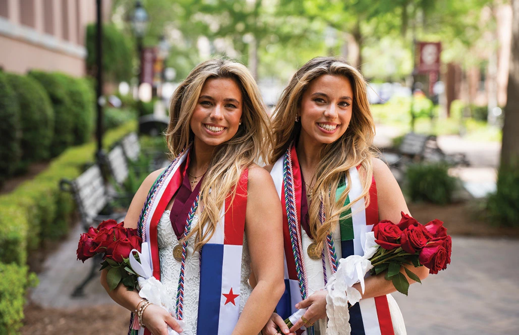 Two graduates smiling with roses and stoles on a tree-lined campus walkway.