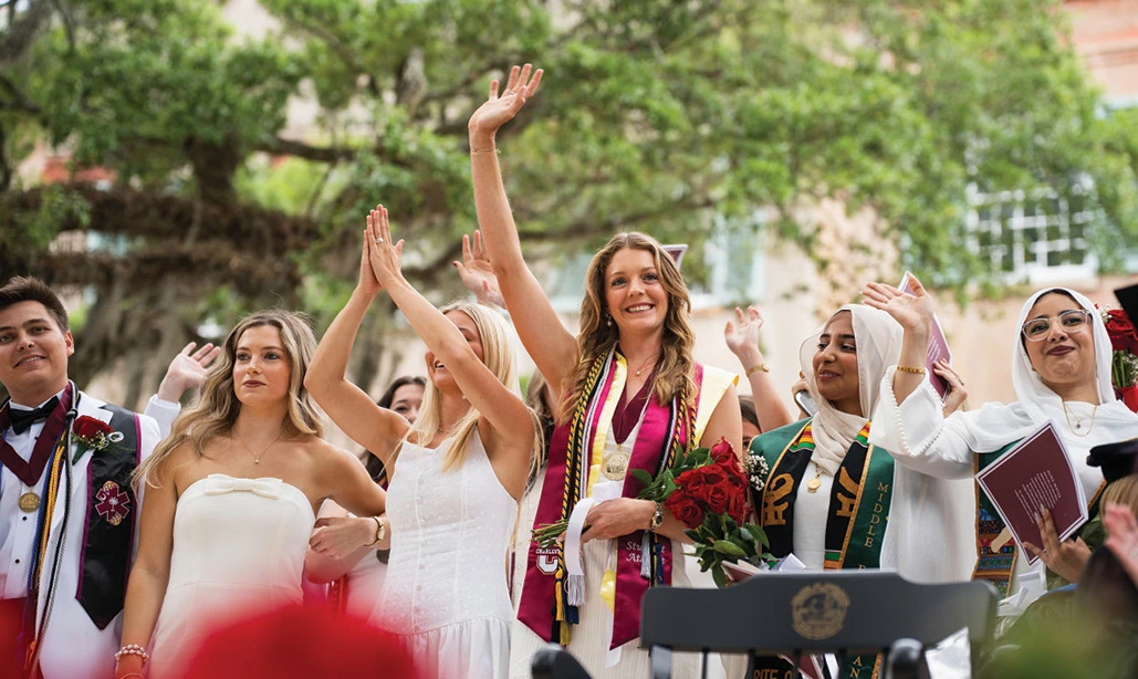 Graduates in white attire waving during an outdoor commencement ceremony.
