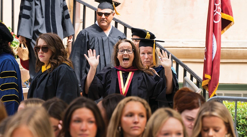 Faculty member in academic regalia waving during a commencement ceremony.