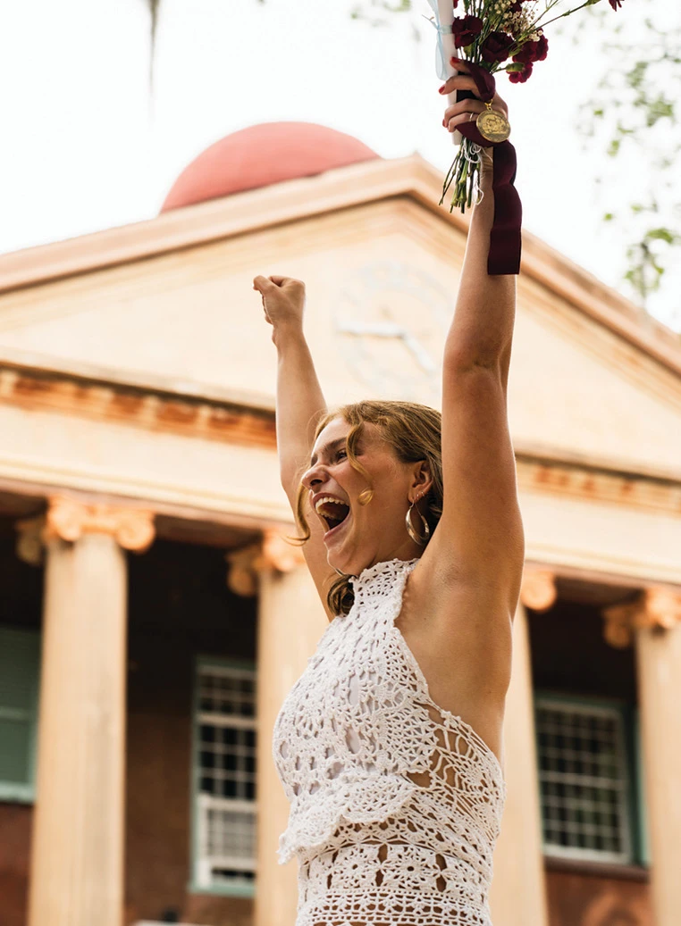 Graduate smiling with raised arms while holding roses in front of a historic campus building.