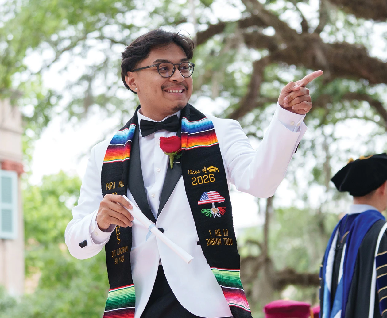 Graduate in a white dinner jacket pointing while holding a diploma during commencement.