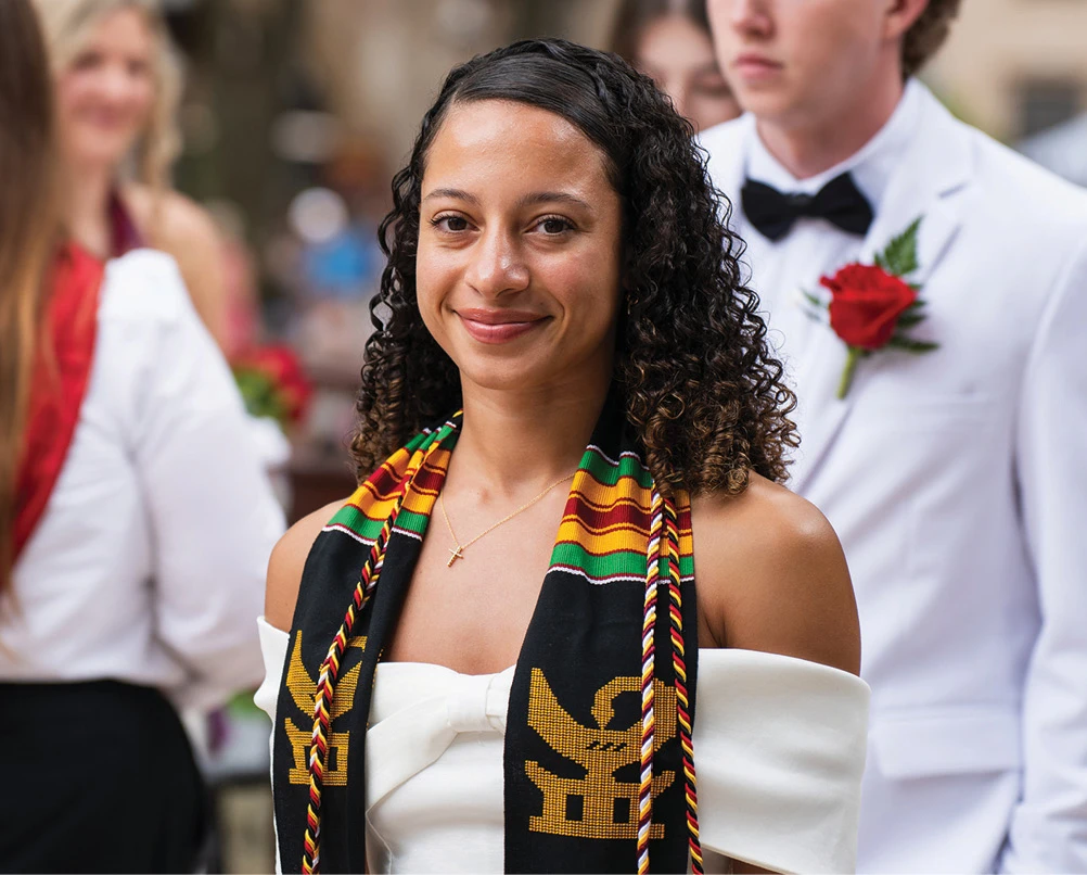 Graduate wearing a Kente stole smiles during an outdoor commencement ceremony.