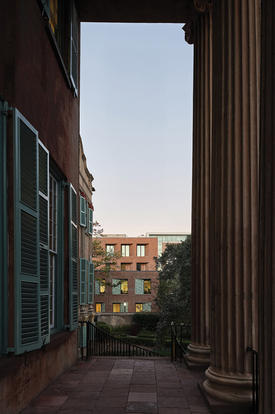 A narrow view between a dark wall with green shutters and a large fluted column. In the distance, a modern brick building with illuminated windows stands against a twilight sky. Paved ground and steps are below.