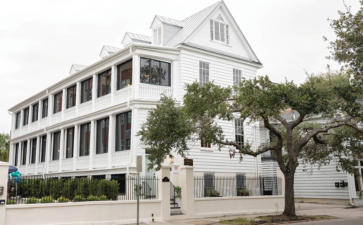 A white, three-story building for the Center for Intentionality, featuring multi-level covered side porches with dark windows. A black wrought-iron fence separates the property from the street. A large tree stands on the right.