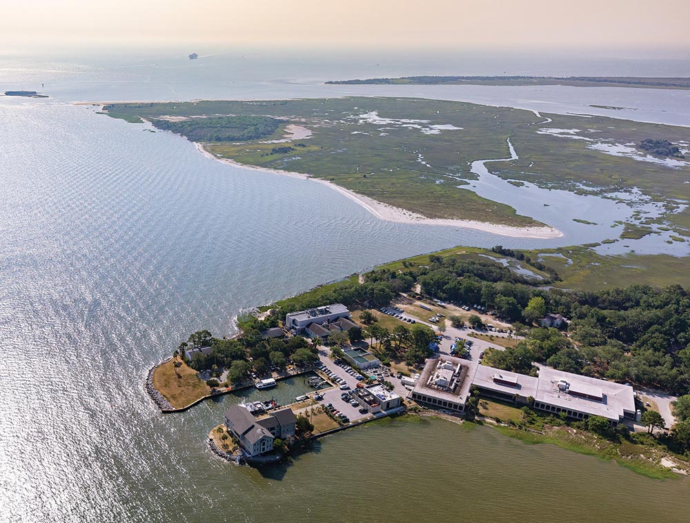 Aerial photograph of a coastal facility on a point of land surrounded by water, with vast green wetlands extending into the distance under a pale sky. The campus includes low-rise buildings, docks, and parked cars.