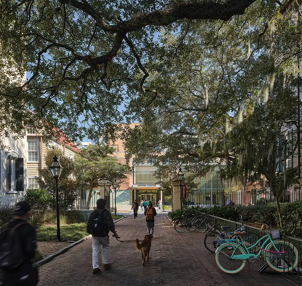 A brick walkway shaded by large live oak trees leads to the entrance of the Simons Center. People stroll along the path; historical buildings with green shutters are on either side. A teal bicycle is parked.