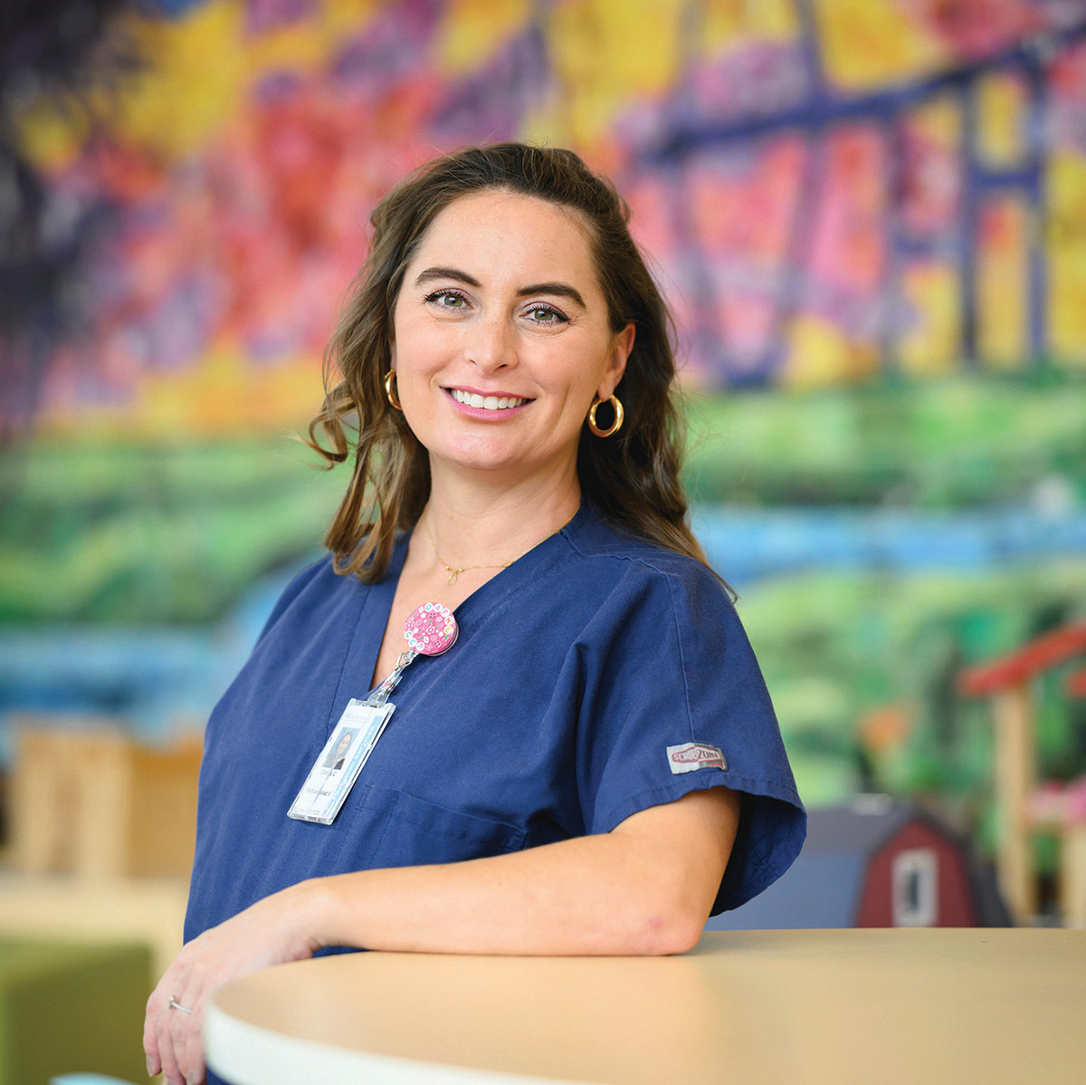 Portrait of a smiling female healthcare professional in blue scrubs leaning against a desk.