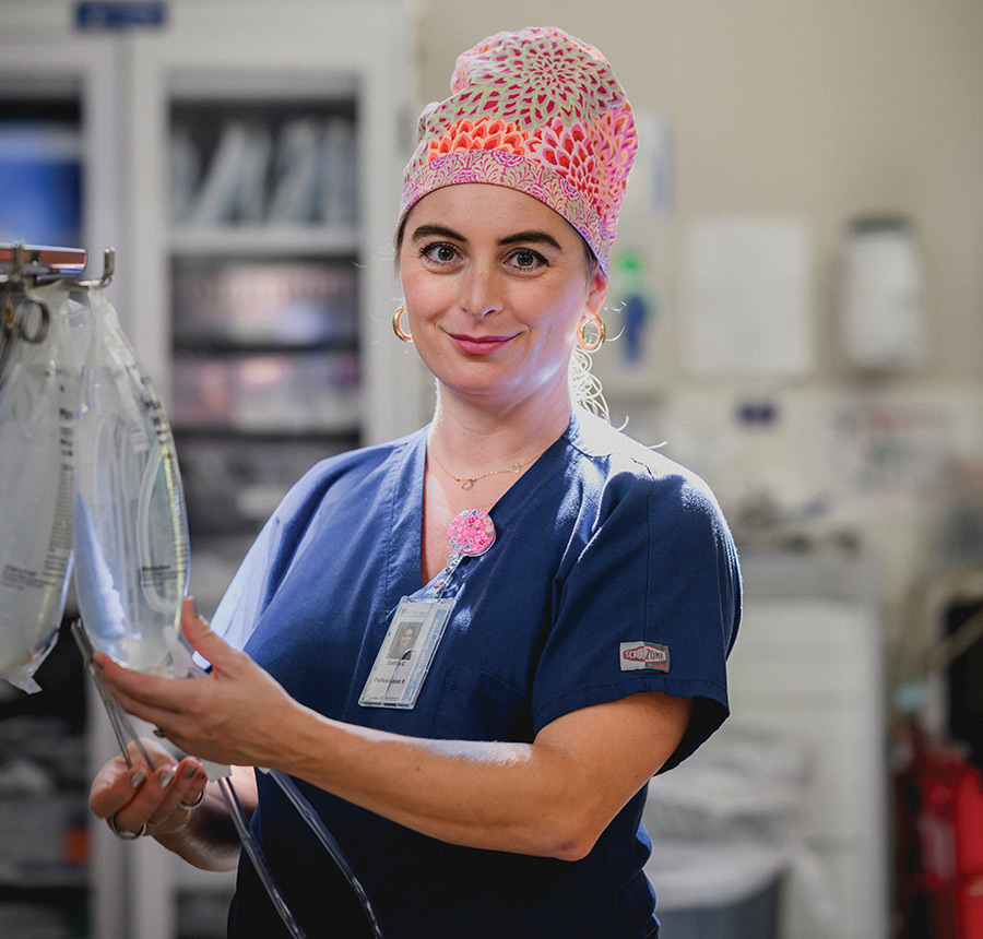 A smiling female perfusionist in scrubs and a colorful surgical cap handling IV fluid bags.