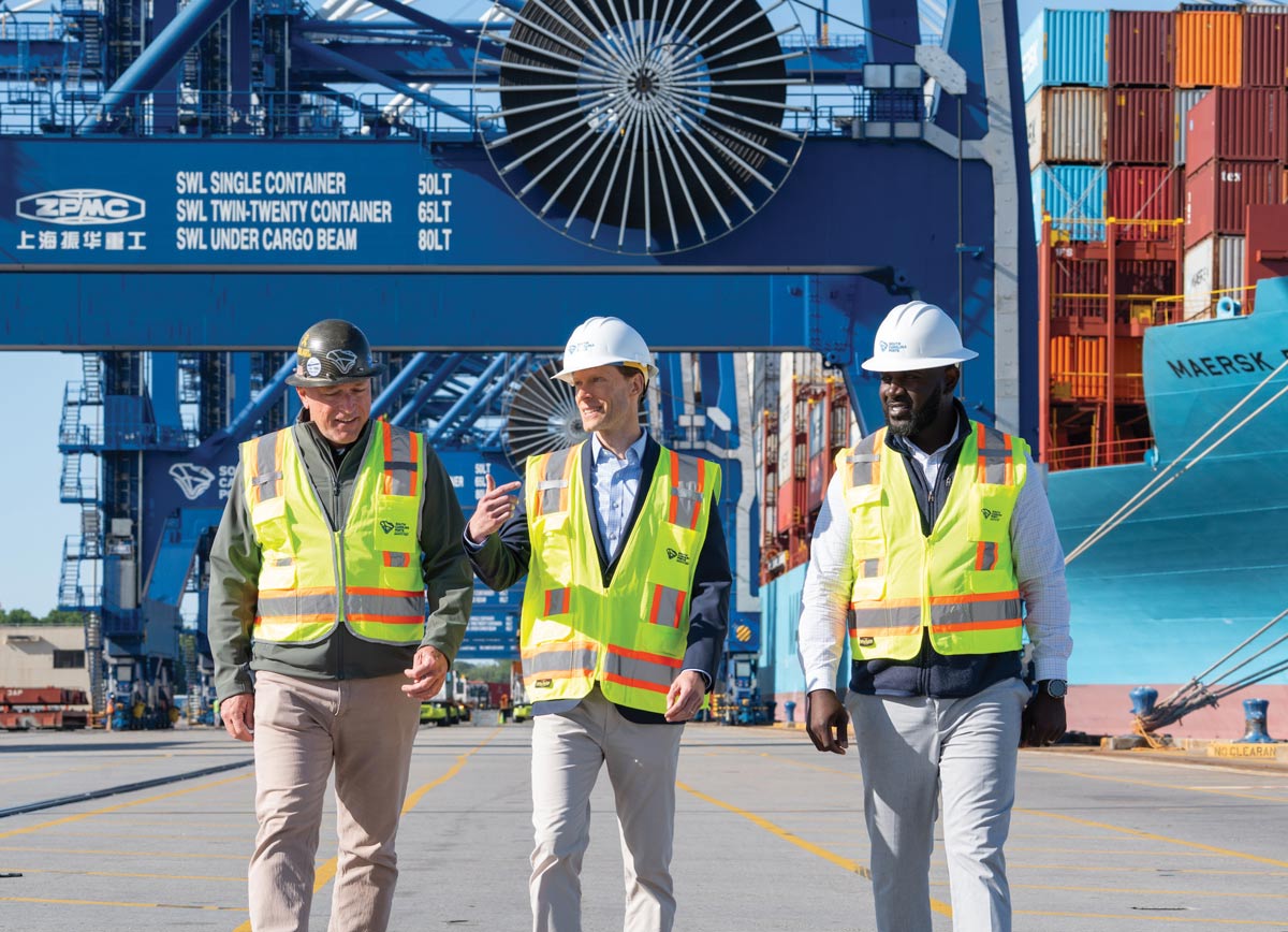 Three port officials in hard hats and high-visibility safety vests walking and talking along a shipping terminal dock.