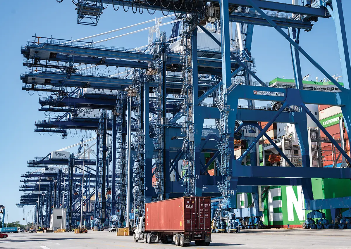 Row of massive blue industrial shipping cranes towering over a semi-truck carrying a red container at a busy port.
