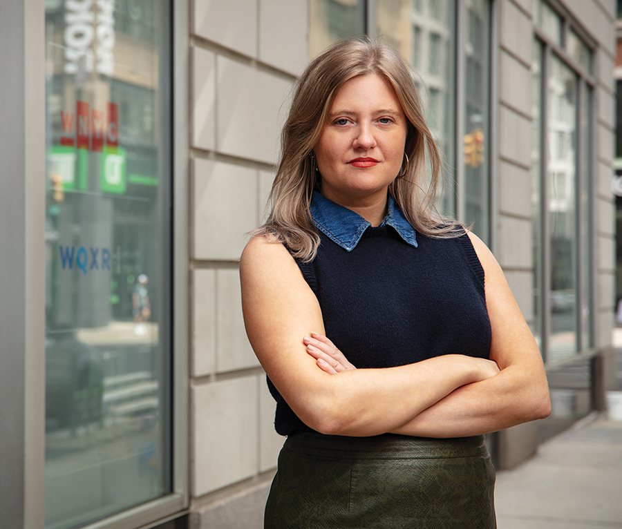 Brooks Brunson, a woman with blonde hair wearing a sleeveless denim-collared top and an olive green textured skirt standing with her arms crossed
