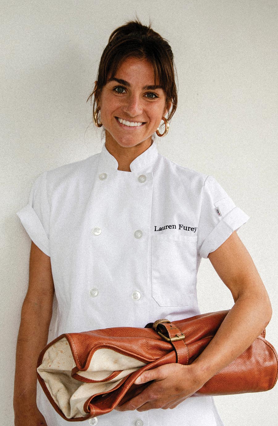 Lauren Furey, a smiling woman with dark hair tied back, wearing a short-sleeved white chef's coat embroidered with her name and gold hoop earrings