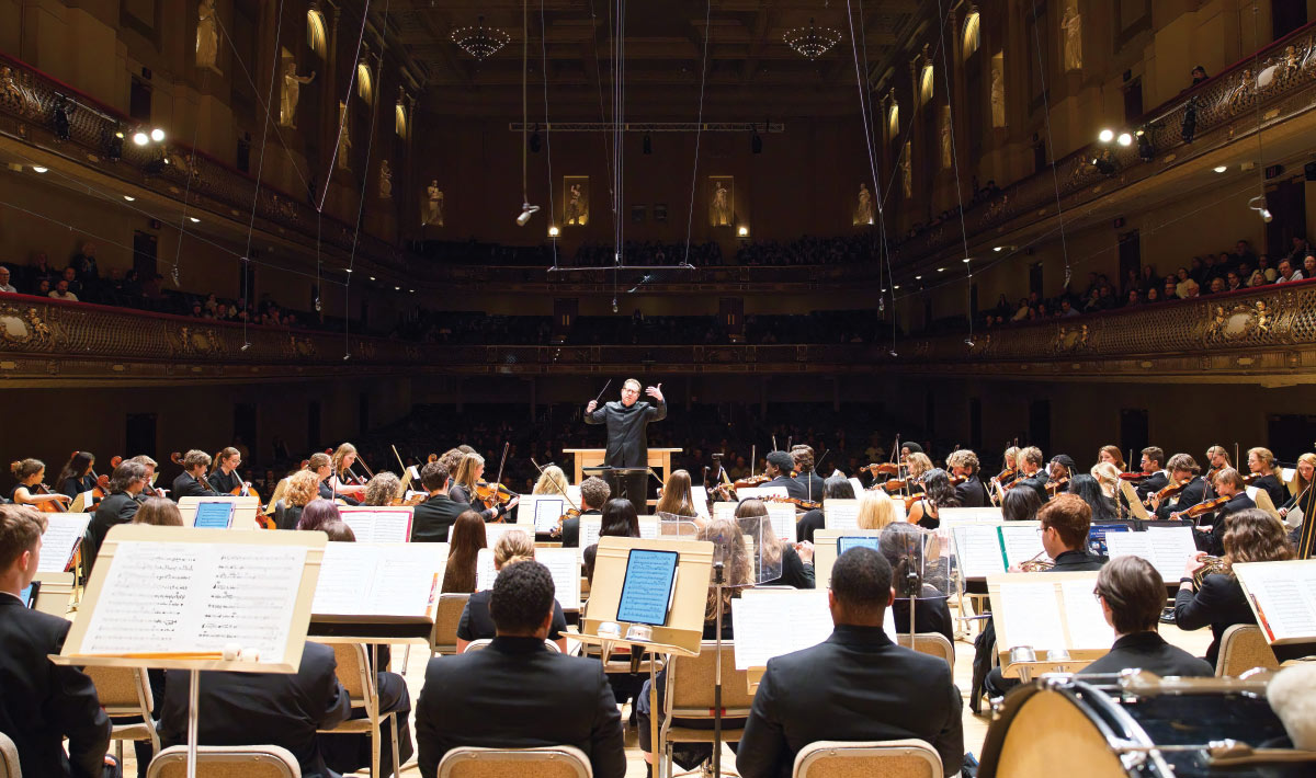 A full symphony orchestra performs on stage under a conductor's baton inside a grand, multi-tiered classical concert hall.