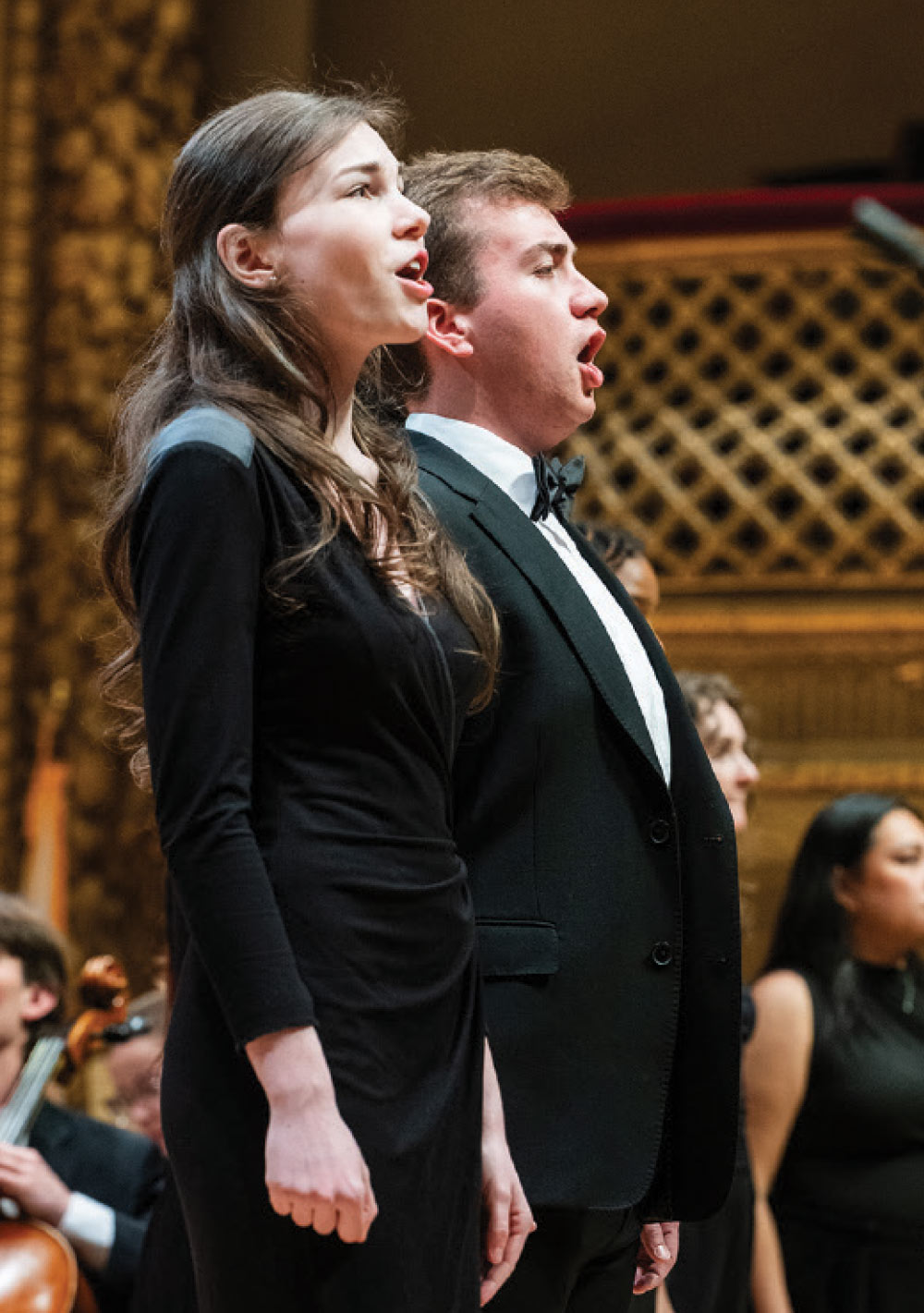 A young man in a tuxedo and a young woman in a black dress sing passionately during a vocal performance in a grand auditorium.