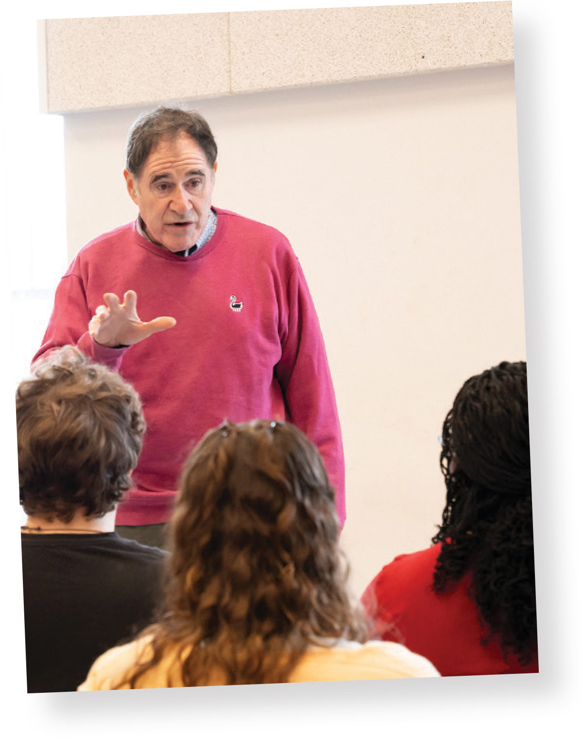 A man wearing a pink sweatshirt gestures with his hand while speaking to a small group of seated students in a classroom.
