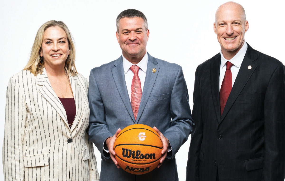 Three professionals stand against a white background, with the center man in a grey suit smiling and holding a Wilson basketball.