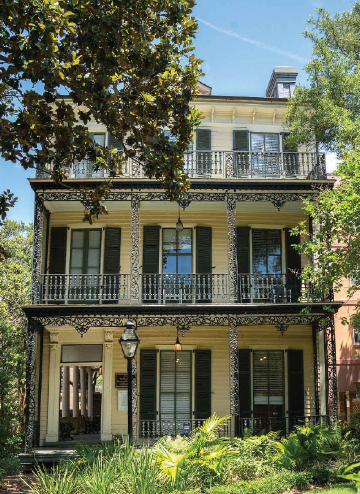 A three-story historic building with yellow siding, dark shutters, and ornate black ironwork balconies, surrounded by lush green foliage.