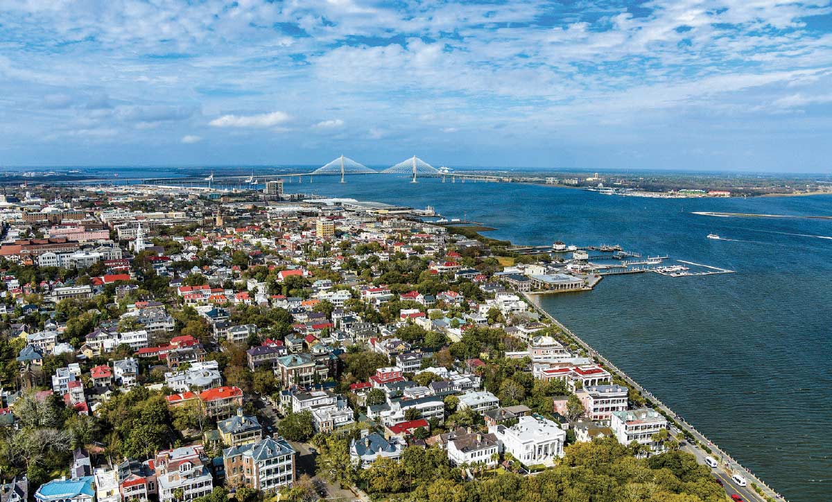 An aerial view of a historic coastal city waterfront showing dense neighborhoods stretching toward a distant large cable-stayed bridge.