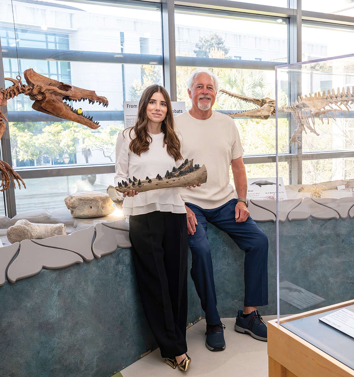 Elizabeth Kane holds a fossilized jawbone alongside Mace Brown inside a museum filled with prehistoric whale skeletons.