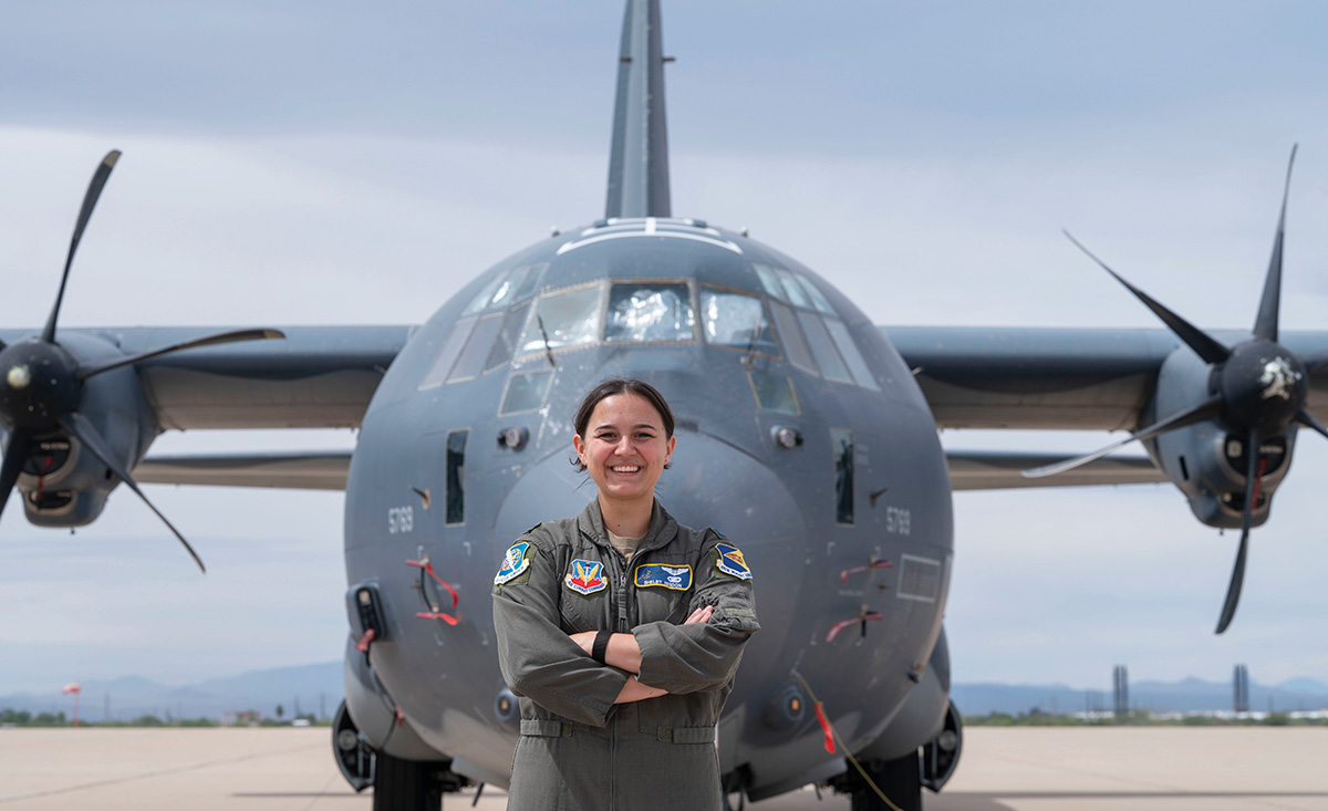 A smiling female pilot in a flight suit stands with arms crossed directly in front of a large, grey military cargo plane.