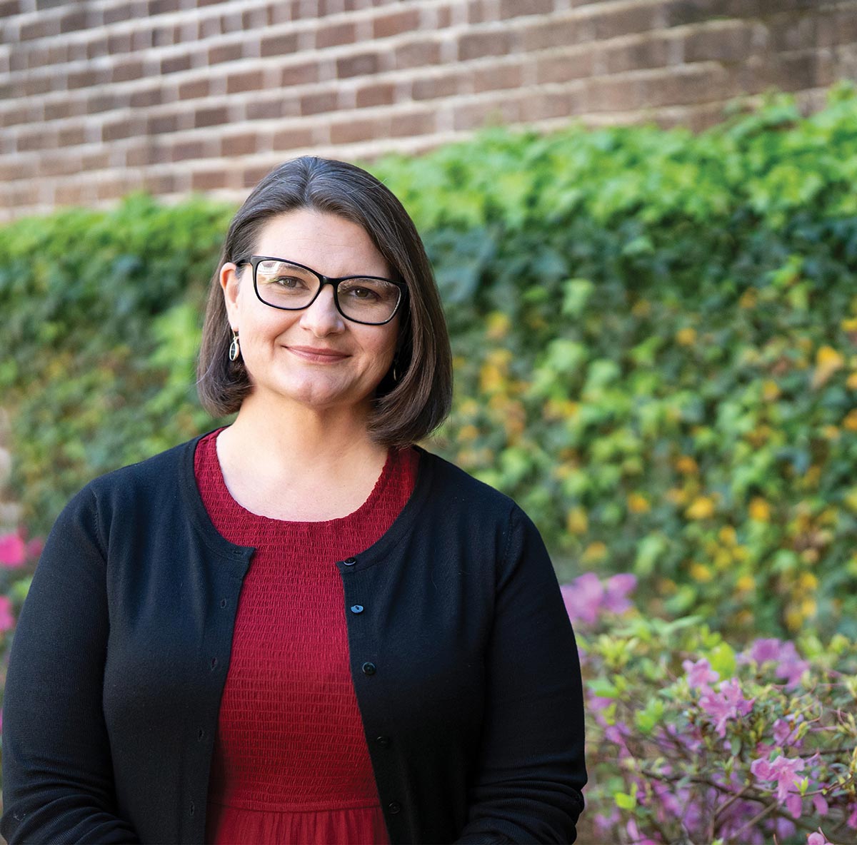 A smiling woman with glasses stands outdoors in front of a brick and ivy wall.