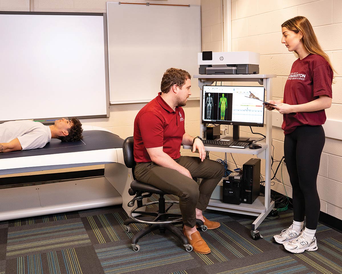 A man and a woman look at a computer screen displaying skeletal and body scan data while a third person lies on an examination table nearby.