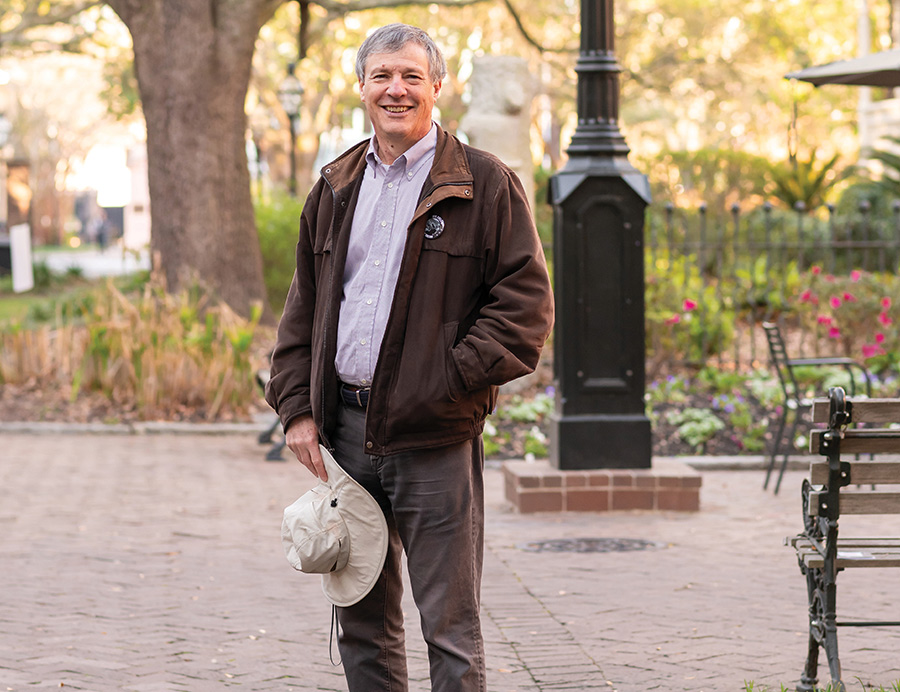 Simon Lewis smiling in a brown jacket stands outdoors in a brick courtyard holding a sun hat.