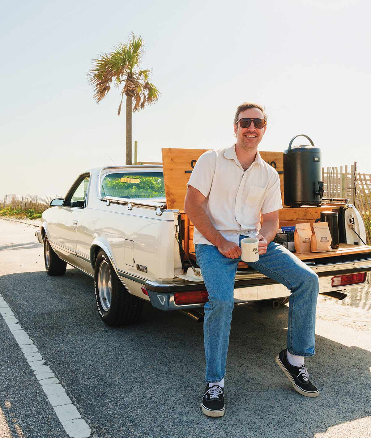 Robert Huffman smiles while sitting on the tailgate of a classic white utility vehicle set up with a mobile coffee station.
