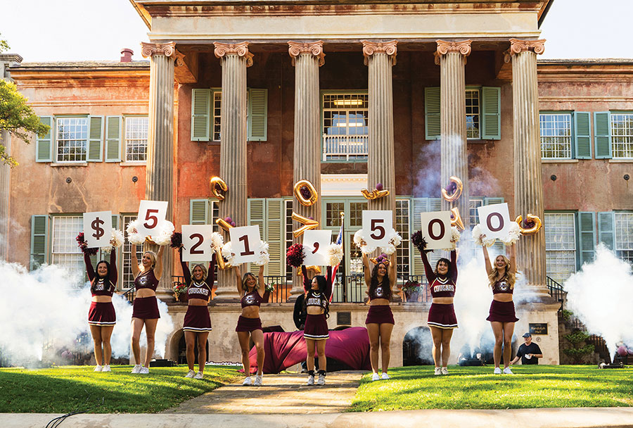 College of Charleston cheerleaders stand outside a historic columned building holding up donation dollar amount signs an outdoor university event.