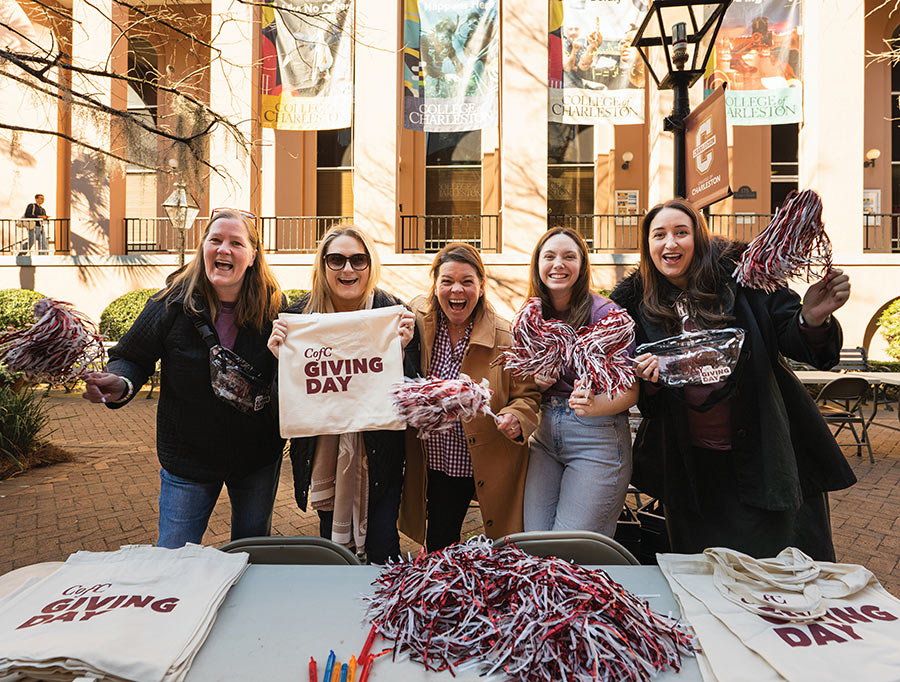 A group of five excited women smile and hold up pom-poms and "CofC Giving Day" tote bags behind a table at an outdoor university event.